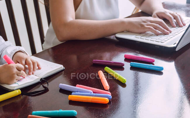 Recorte irreconocible mujer portátil de navegación mientras que el niño pequeño sentado en la mesa y el dibujo con marcadores en el cuaderno - foto de stock