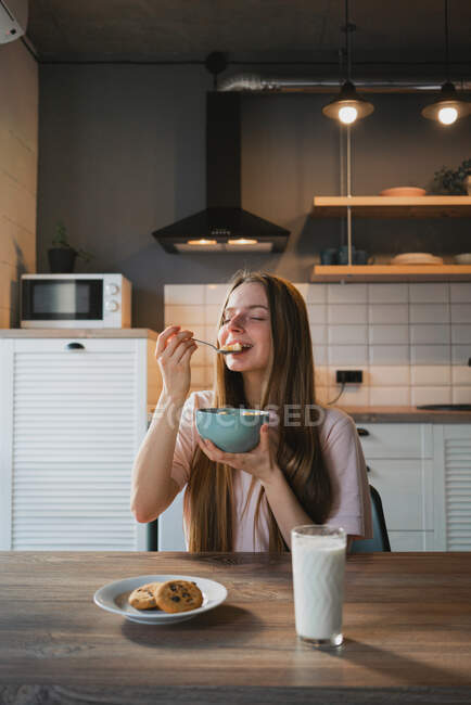 Jeune femelle avec cuillère et bol profitant de savoureux anneaux de maïs dans la cuisine — Photo de stock