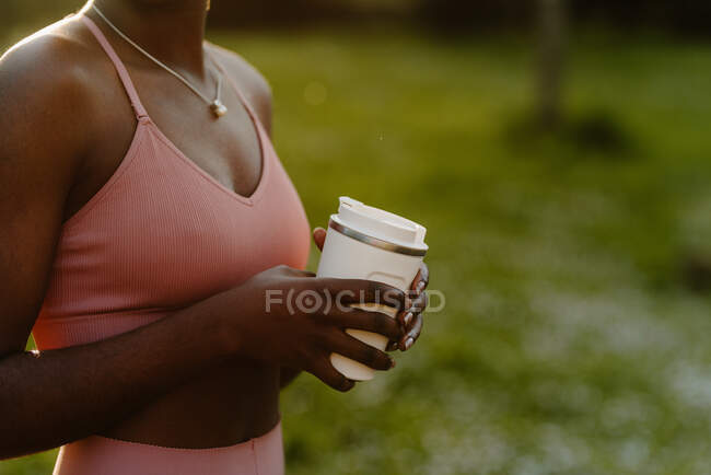 Crop African American female athlete in activewear standing with cup of beverage in park in evening and looking away — Stock Photo