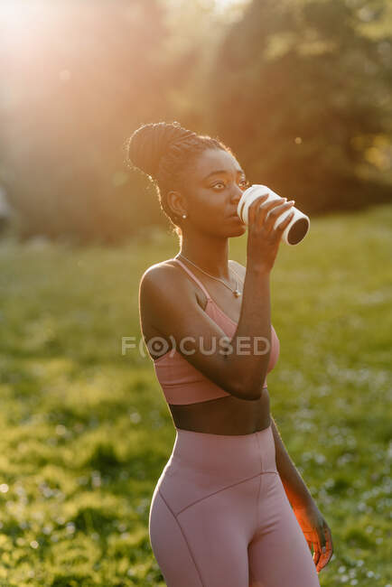 Возбужденная афроамериканка-атлет в активной одежде, стоящая с чашкой напитка в парке вечером и отворачивающаяся — стоковое фото