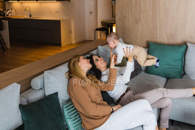 Alegre pareja de mujeres homosexuales jugando con lindo niño mientras se divierten y disfrutan de fin de semana juntos en casa - foto de stock