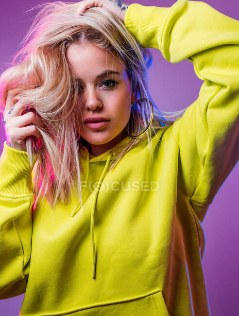 Determined female in yellow hoodie touching blond hair while standing on purple background in studio with blue and pink neon lights — Stock Photo