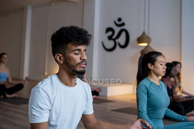 African American man with group of diverse people sitting in Lotus pose with closed eyes and mediating while practicing yoga together during class in studio — Stock Photo