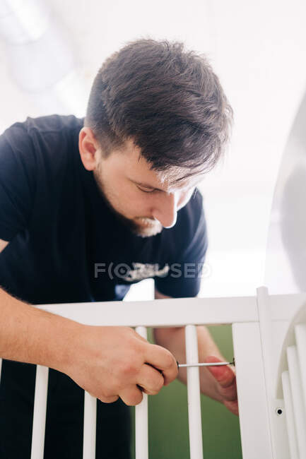 From below of bearded male with screwdriver leaning forward while mounting crib at home on sunny day — Stock Photo