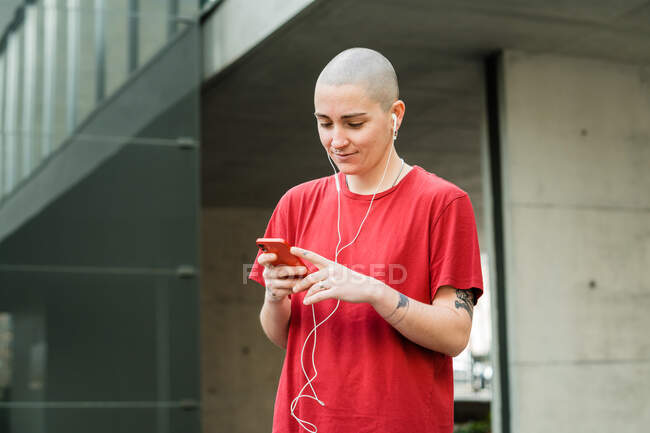 Junge glückliche homosexuelle Frau in T-Shirt und Kopfhörer mit Handy, die auf den Bildschirm schaut, während sie Musik hört — Stockfoto