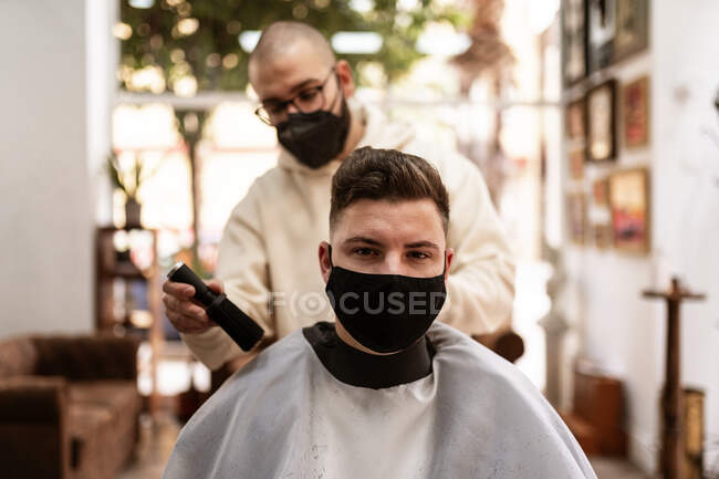 Мужчина парикмахер в тканевой маске для лица с щеткой для удаления волос с плаща клиента в парикмахерской — стоковое фото
