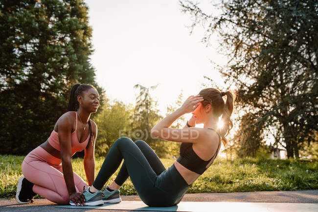 Side view of slim female athlete doing abdominal crunches with help of black sportswoman during training at sunset in park — Stock Photo