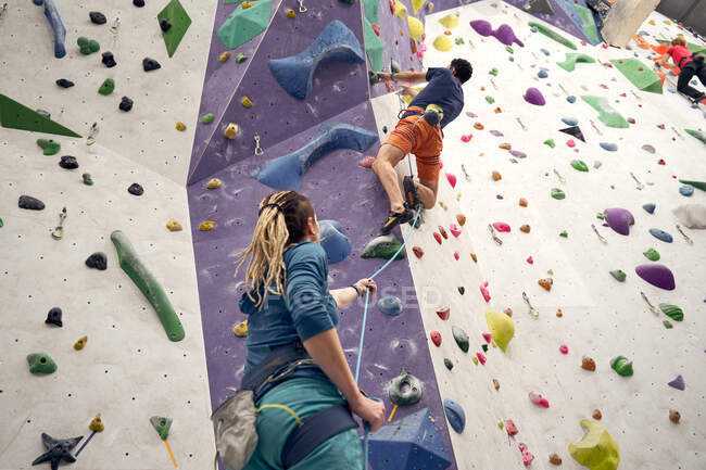 Visão traseira de baixo ângulo do instrutor fêmea que apoia o alpinista masculino no arnês que escala a parede artificial no centro de pedregulho — Fotografia de Stock