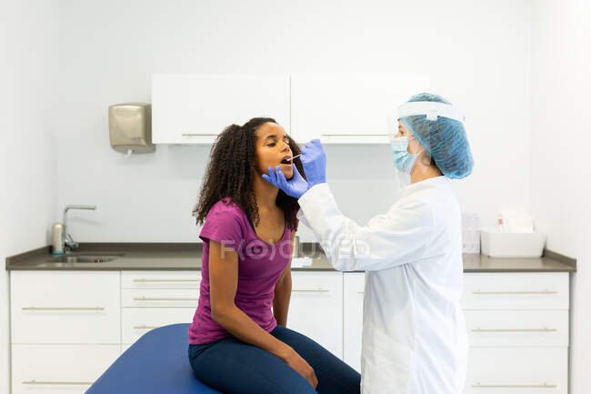 Female medical specialist in protective uniform, latex gloves and face mask doing mouth swab test with cotton bud on African American female patient in clinic during coronavirus outbreak — Stock Photo