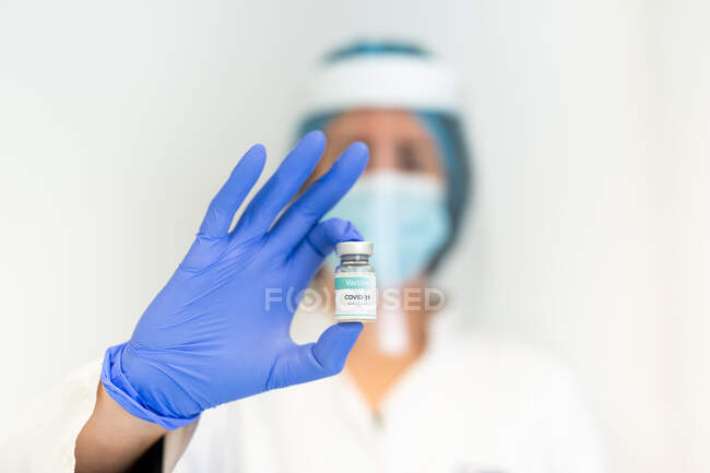 Doctora en guantes de látex y protector facial de pie con líquido químico en un vial de vidrio que se prepara para vacunar al paciente en la clínica durante el brote de coronavirus - foto de stock