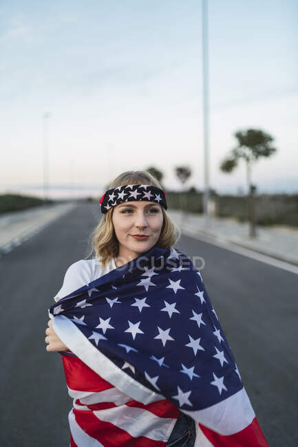 Delighted American female standing wrapped with national USA flag on roadway at sunset and looking away — Stock Photo