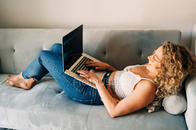 Вид на молодую самозанятую женщину в повседневной одежде и очках лежащую на удобном диване и работающую дистанционно на ноутбуке — стоковое фото