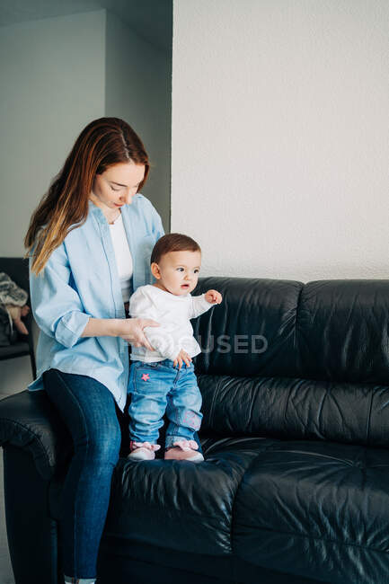 Орієнтована молода мати в повсякденному одязі навчає чарівну дитину ходити на затишному дивані вдома — стокове фото