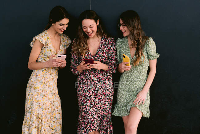 Content young best female friends in trendy apparel with cellphones standing on urban walkway against wall — Stock Photo
