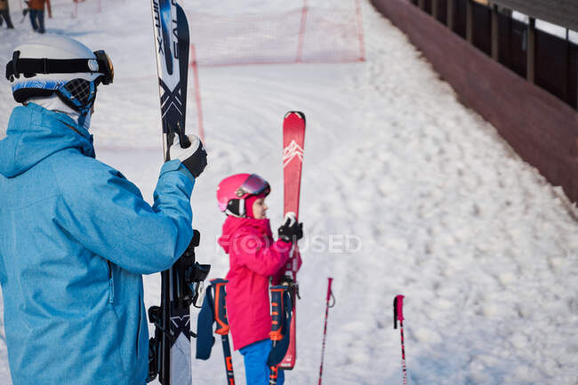 Apto para padres y niños con ropa deportiva cálida y cascos que se ponen esquís mientras están de pie en la colina nevada en el suburbio en el clima soleado de invierno - foto de stock