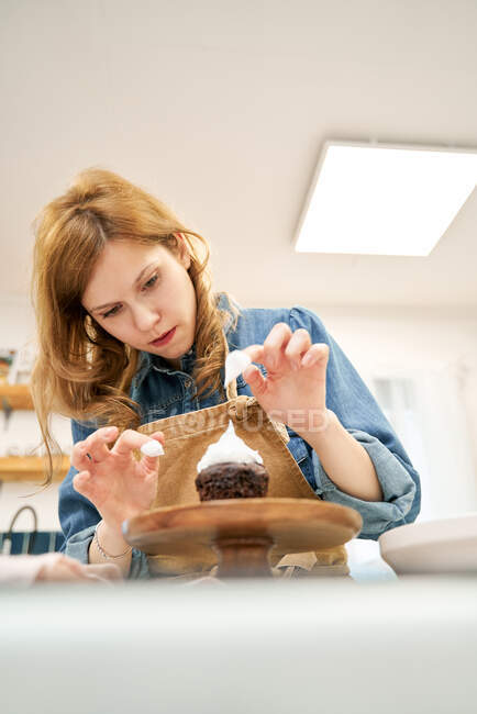 De baixo de jovem fêmea decorando cupcake de chocolate com chantilly doce no carrinho de bolo enquanto cozinha em casa — Fotografia de Stock