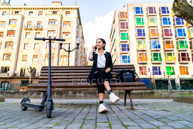 Зміст молода етнічна жінка-підприємець сидить на лавці, виступаючи на смартфоні, дивлячись далеко від електричного скутера та міських будівель — стокове фото
