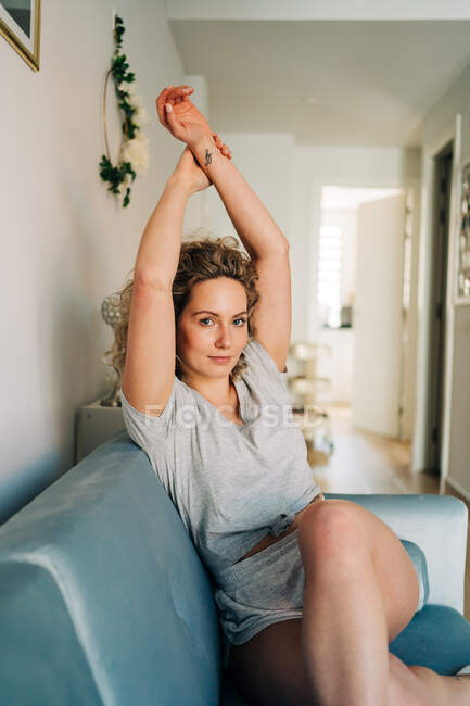 Mujer atractiva positiva en pantalones cortos sentados en un acogedor sofá en la sala de estar estirándose con los brazos hacia arriba y mirando a la cámara - foto de stock