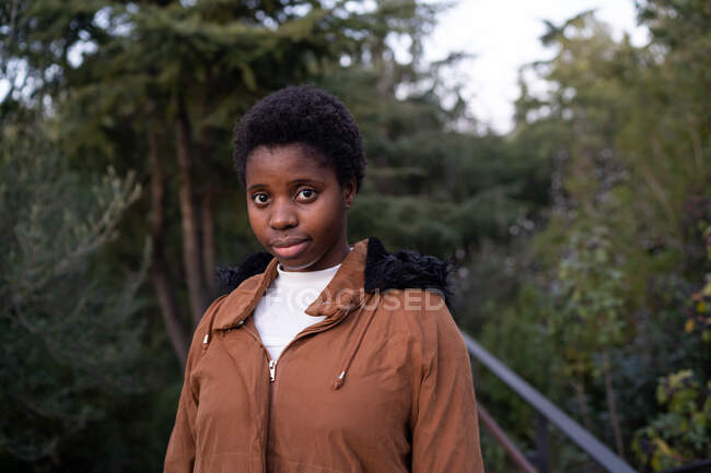 Junge, ruhige Afroamerikanerin in warmer Jacke steht im grünen, üppigen Park und blickt an einem Frühlingstag in die Kamera — Stockfoto