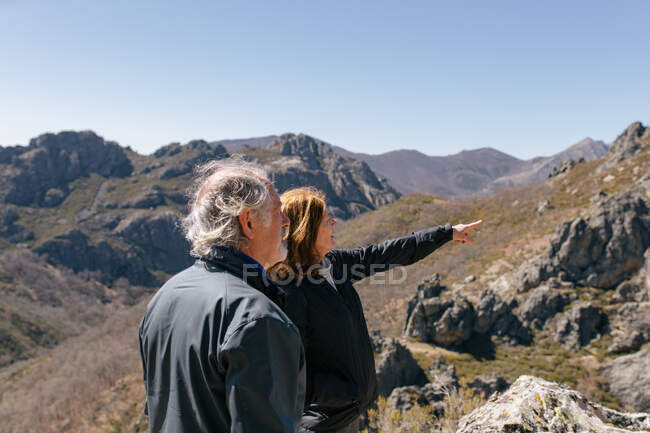 Anciano con mujer apuntando lejos disfrutando de la naturaleza de las montañas mientras caminan juntos - foto de stock