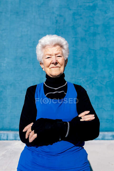 Урожай счастливой зрелой женщины в спортивной одежде, стоящей напротив синей стены в открытом спортивном центре и смотрящей в камеру с улыбкой и скрещенными руками — стоковое фото