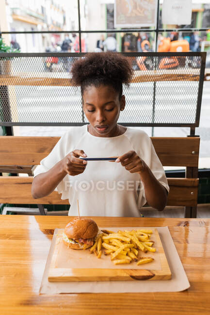 Frontansicht einer ruhigen Afroamerikanerin in stilvoller Kleidung, die Fotos von leckeren Burgern und Pommes frites macht, die auf einem hohen Tisch im Fast-Food-Restaurant serviert werden. — Stockfoto