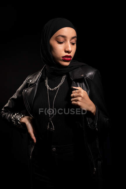 Привлекательная молодая исламская женщина в черной одежде в кожаной куртке и хиджабе, нежно смотрящая на черную студию — стоковое фото