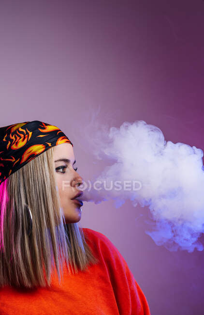 Vue latérale de la femelle cool dans la tenue de style de rue fumer e cigarette et expirer la fumée par le nez sur fond violet en studio avec éclairage au néon rose — Photo de stock