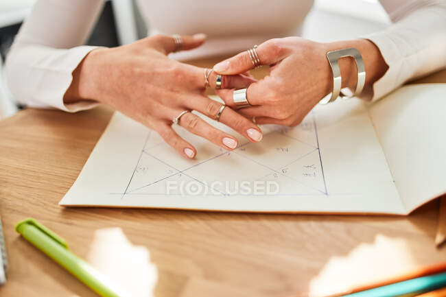 Cosecha irreconocible astróloga femenina en anillos y pulseras que trabajan en el escritorio con álbum de papel en casa - foto de stock