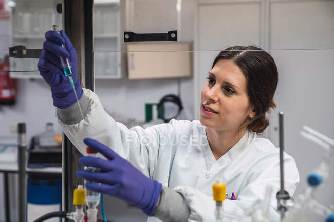 Урожай концентрована жінка-вчена в білому халаті і рукавичках, що проводить хімічний експеримент з речовиною і шприцом під час роботи в сучасній лабораторії — стокове фото