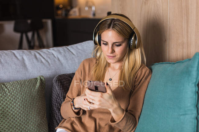Молода жінка сидить на дивані і насолоджується музикою в навушниках, дивлячись на екран смартфона — стокове фото
