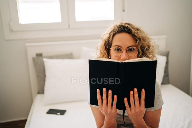 Positive young female with curly blond hair in panties and eyeglasses smiling while sitting on cozy bed and reading interesting book — Fotografia de Stock