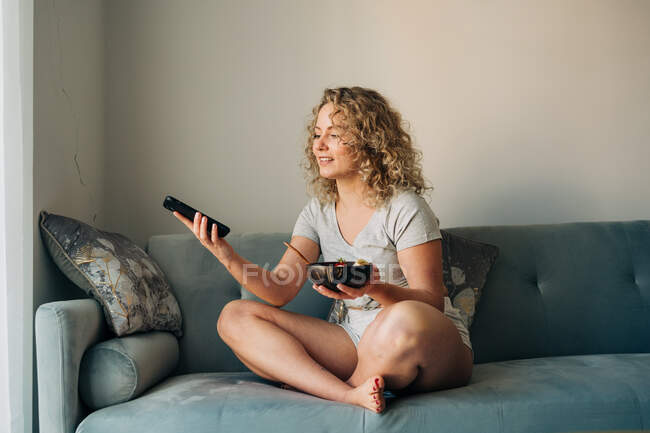 Fröhliche junge Frau in kurzen Hosen am Handy, während sie mit gekreuzten Beinen auf der Couch sitzt und eine Schüssel mit leckerem Haferbrei in der Hand hält — Stockfoto