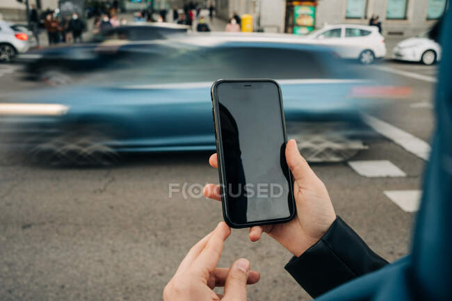 Recorte mujer anónima de pie cerca de la carretera con tráfico ocupado y la navegación del teléfono móvil con pantalla en negro - foto de stock