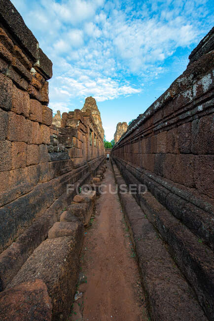 Anonymous female tourist on narrow walkway between aged masonry walls of temple complex in Cambodia under cloudy blue sky — Stock Photo