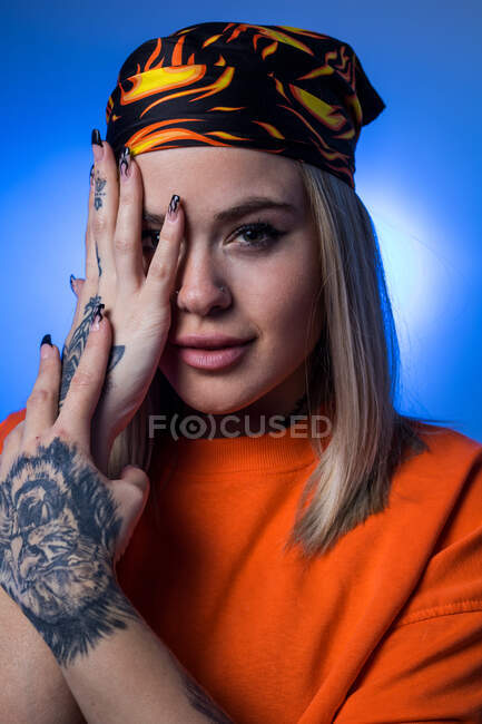 Sonriente mujer elegante en bandana y con tatuajes que cubren la mitad de la cara con la mano y mirando a la cámara sobre fondo azul en el estudio - foto de stock