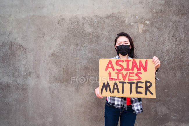 Donna etnica in maschera e con cartello di cartone con iscrizione Asian Lives Matter che protesta in strada e guarda la macchina fotografica — Foto stock