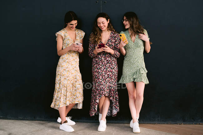 Содержание молодых лучших подруг в модной одежде с сотовыми телефонами, стоящими на городской дорожке напротив стены — стоковое фото