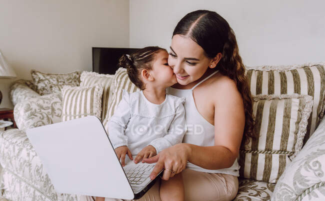 Ethnic woman with little girl on knees sitting in sofa at home and watching cartoon on netbook together — Stock Photo