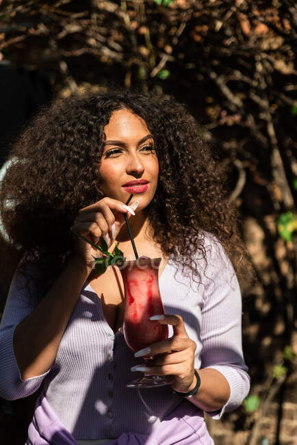 Joven hembra étnica positiva usando atuendo casual disfrutando de cóctel dulce fresco y mirando hacia otro lado mientras está de pie en el jardín soleado - foto de stock