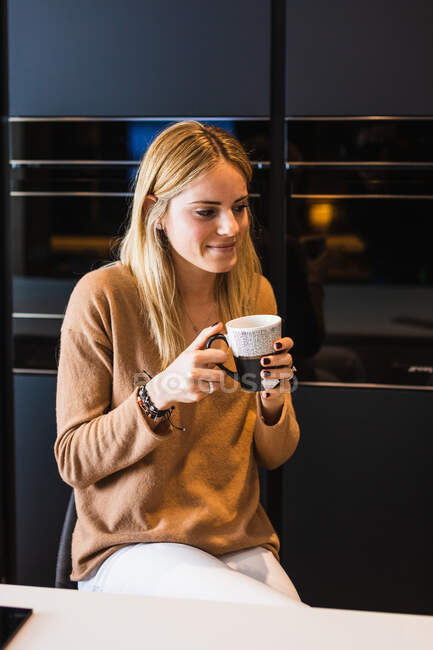 Content female with mug of hot beverage sitting on stool on the counter in kitchen at home looking away — Stock Photo