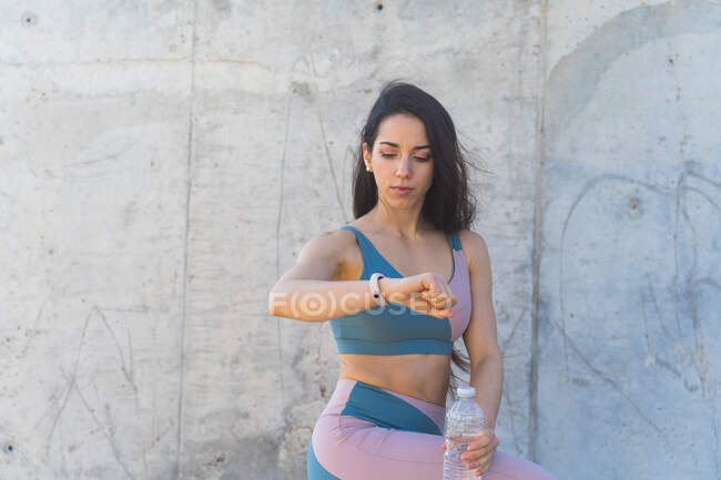 Young fit female athlete in sportswear watching heartbeat on wearable bracelet during break from workout on gray background outdoors — Photo de stock