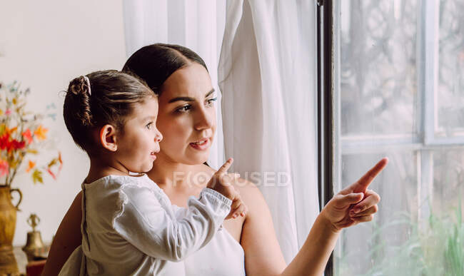 Seitenansicht der ethnischen Mutter mit ihrer niedlichen kleinen Tochter, die aus dem Fenster schaut, während sie zu Hause im Zimmer steht — Stockfoto