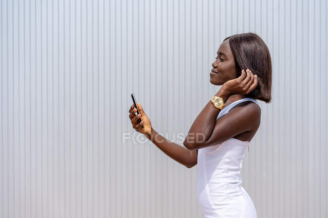 Positivo elegante donna afroamericana in abito bianco navigando moderno telefono cellulare mentre in piedi vicino muro edificio sulla strada soleggiata — Foto stock