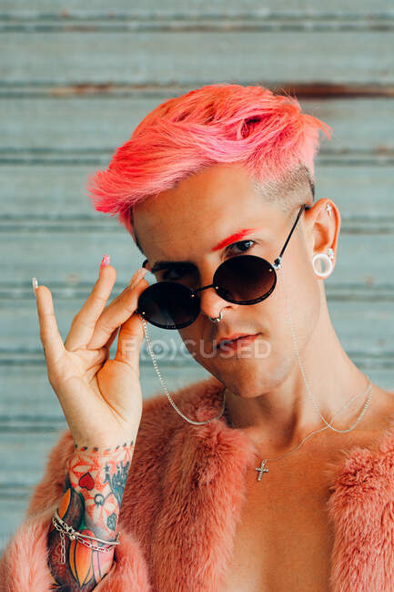 Joven gay en elegante desgaste con tatuaje, pelo rosa y manicura con chaqueta esponjosa sosteniendo gafas de sol mientras mira a la cámara - foto de stock