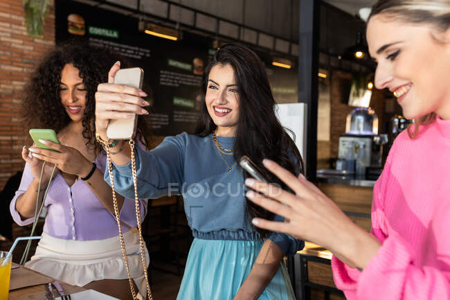 Sorrindo jovens amigas vestindo roupas casuais tirando selfie e navegando telefones celulares enquanto se reúnem para o almoço no restaurante — Fotografia de Stock