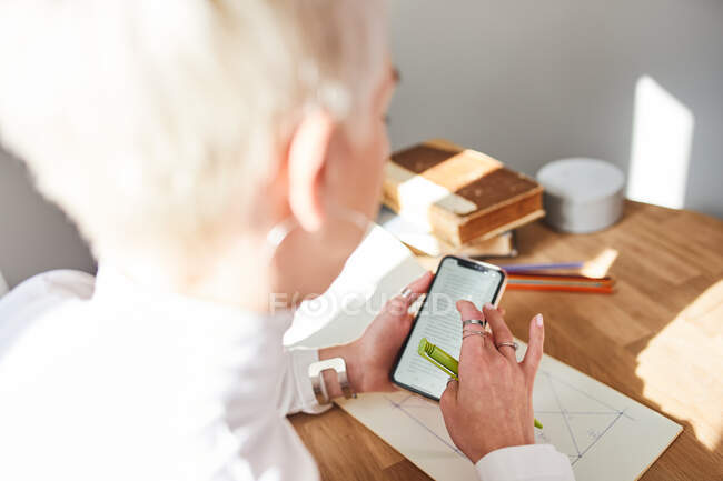 Vue arrière d'une astrologue anonyme avec des cheveux courts et un téléphone portable regardant l'écran à la maison — Photo de stock