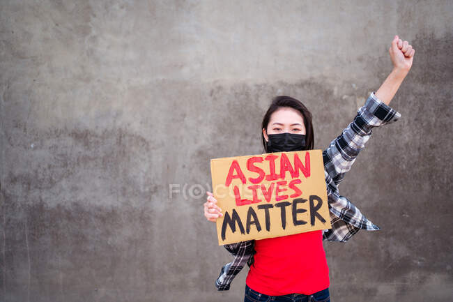 Етнічна жінка в масці і з картонним плакатом з написом азійське життя Протестують з піднятою рукою на вулицях міста і дивляться на камеру — стокове фото