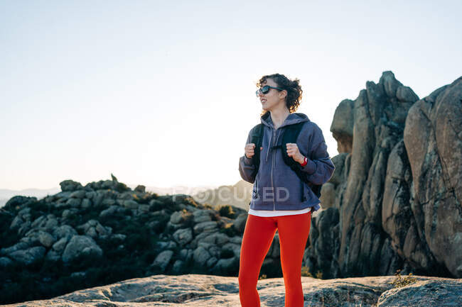 Allegro giovane esploratrice donna con cappuccio e occhiali da sole sorridente mentre ammira la natura durante il trekking nella valle rocciosa montuosa nella giornata di sole — Foto stock