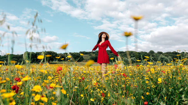 Happy female in red sundress, hat and handbag standing looking up on blossoming field with yellow and red flowers with outstretched arms enjoying on warm spring summer day — Stock Photo